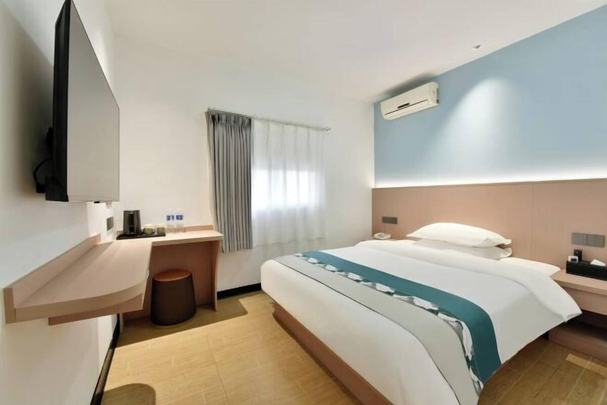 A hotel room with a bed, bedside table, TV, air conditioning unit, and a window with curtains.