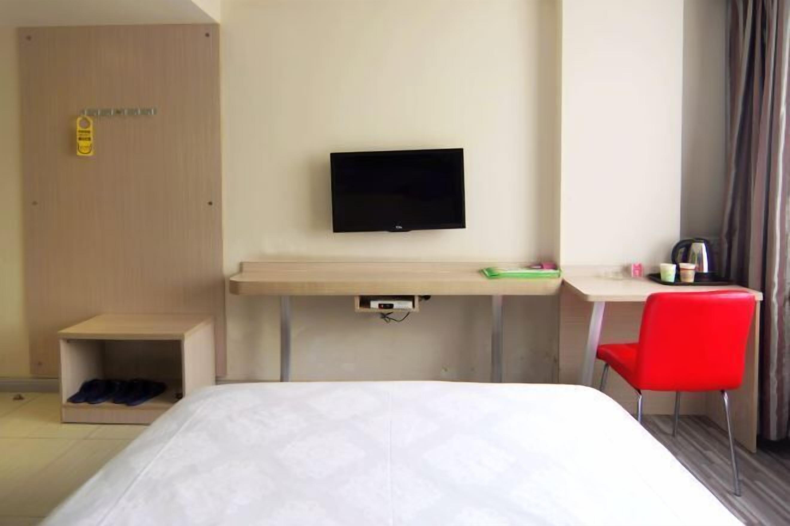 A hotel room with a bed, a desk with a flat-screen TV, a red chair, and a wardrobe.