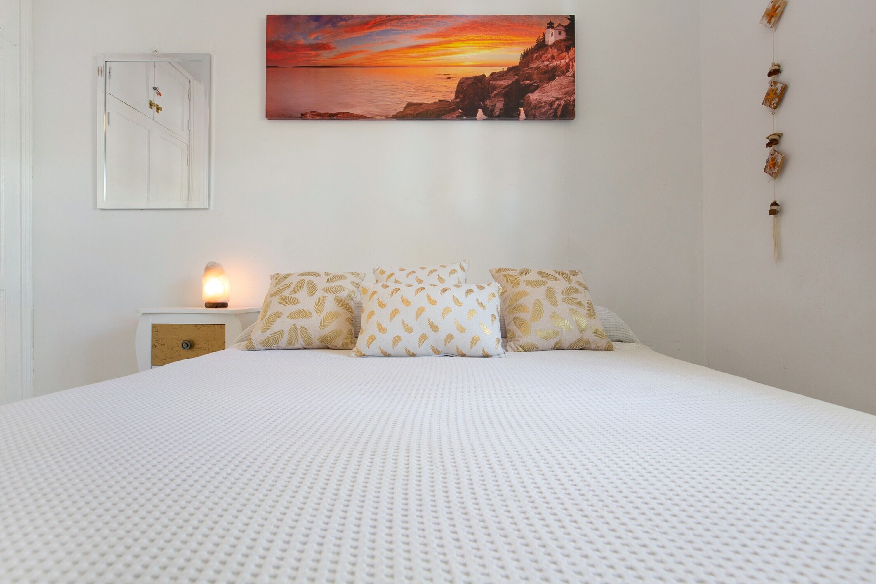 Une chambre à coucher avec un grand lit, une table de chevet avec une lampe, un tableau représentant un coucher de soleil et un miroir fixé au mur.