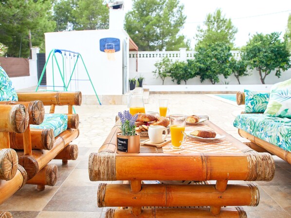 A bamboo furniture set with a table, chairs, and a sofa, a glass pitcher, a mug, and croissants on a checkered tablecloth.