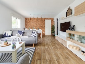 A modern living room with a grey sofa, a dining area with a brick wall, and a TV mounted on a wooden shelf.