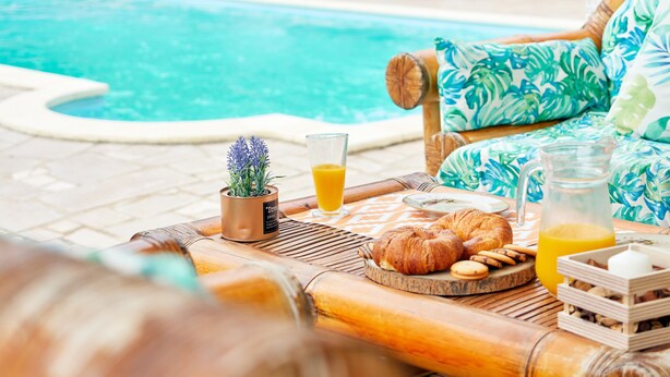 A poolside breakfast setup with a table, a chair, a small plant, and a glass of orange juice.