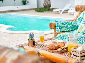A poolside breakfast setup with a table, a chair, a small plant, and a glass of orange juice.