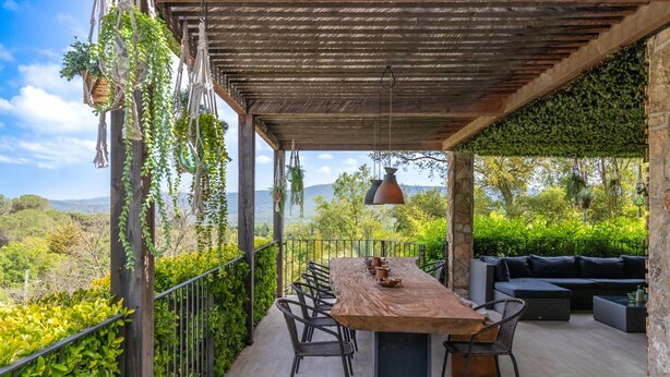 A covered outdoor dining area with a long wooden table and chairs, a hanging pendant light, and a stone wall.
