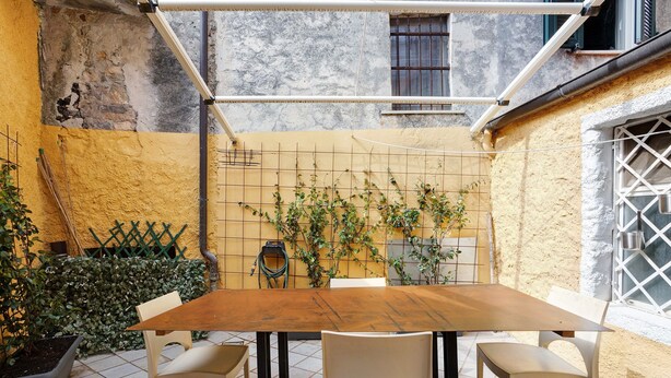 Een terras met een houten tafel en vier stoelen, omgeven door een gele muur met rasterpatroon en een groene klimplant.