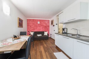 Une cuisine de taille réduite avec des meubles blancs, une table à manger en bois et un canapé à l’arrière-plan.