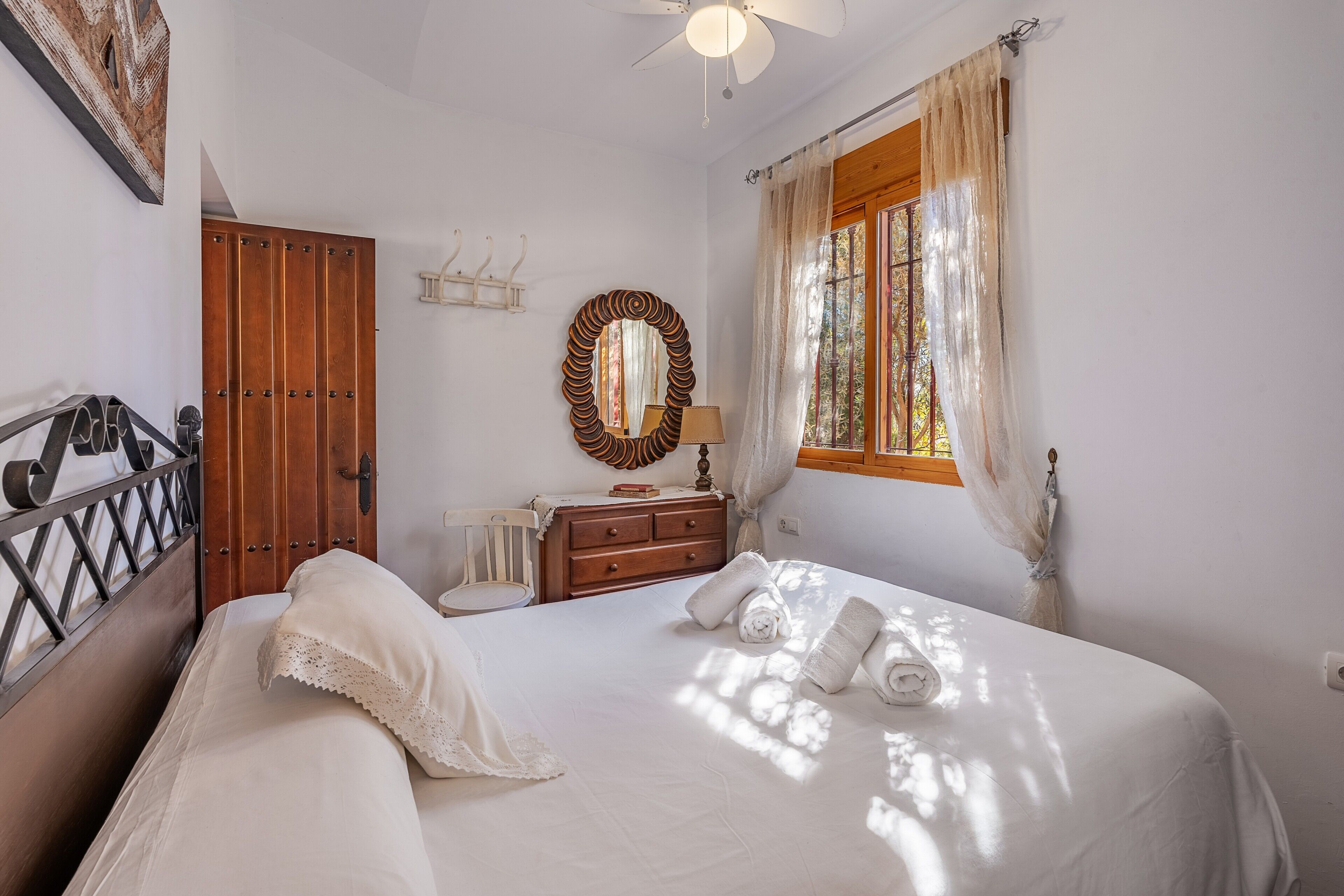 Una camera da letto con un letto, una porta in legno, uno specchio, una finestra con tende e una ventola a soffitto.