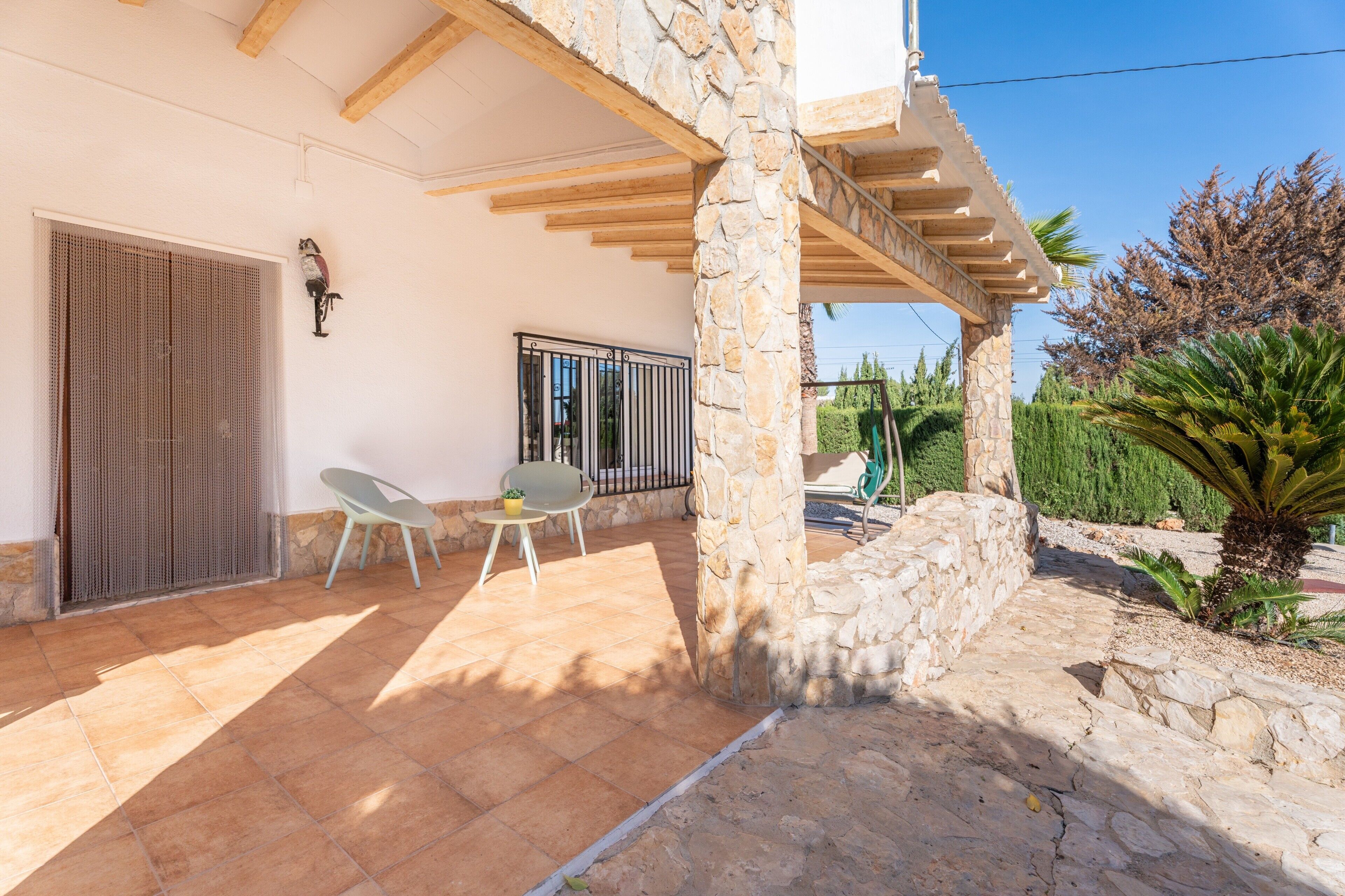 Un patio cubierto con pilares de piedra, una reja metálica y una pequeña mesa con una silla.