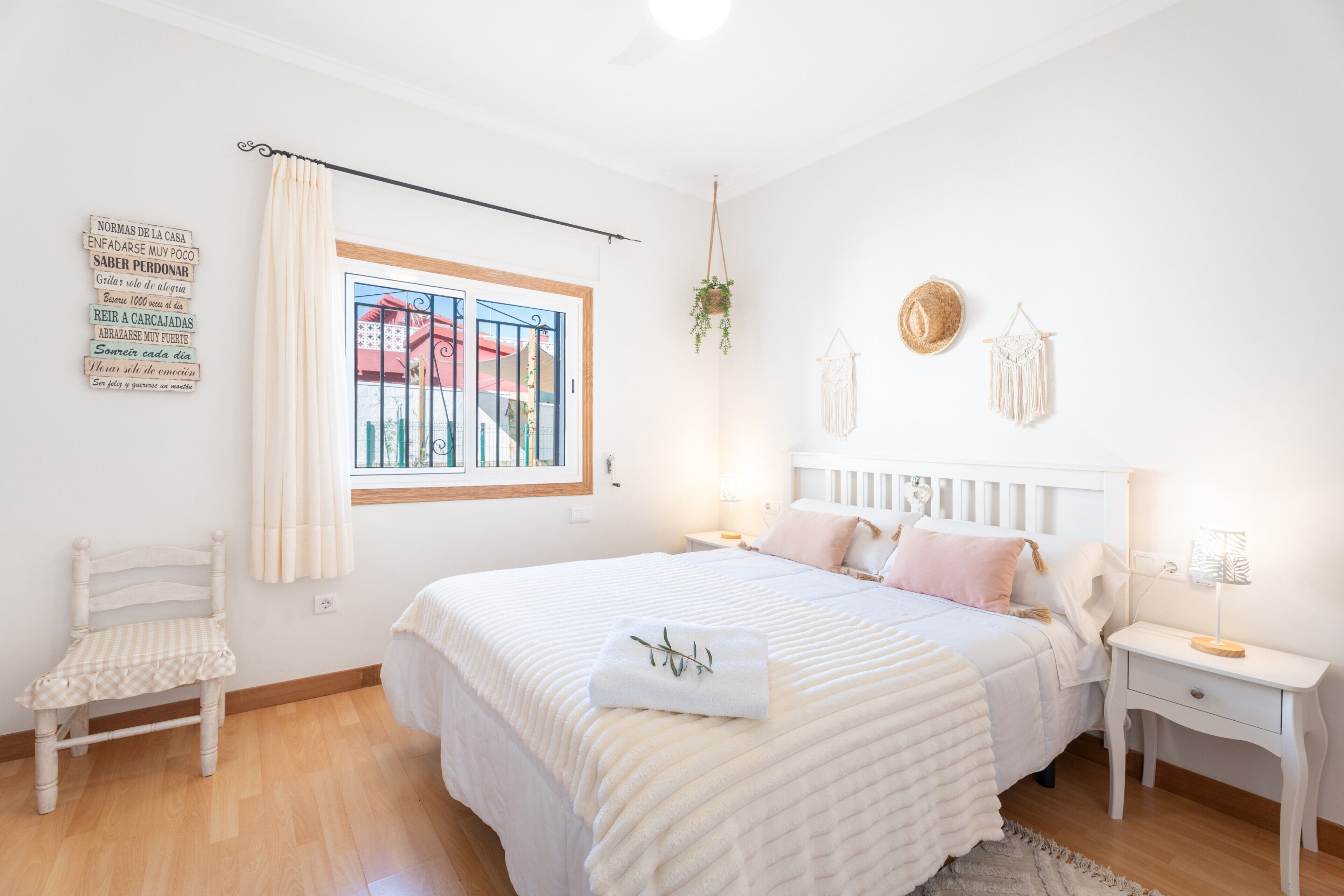 Un dormitorio con cama, una silla, una mesita de noche, una lámpara, una planta colgante y una ventana con vista a edificios.