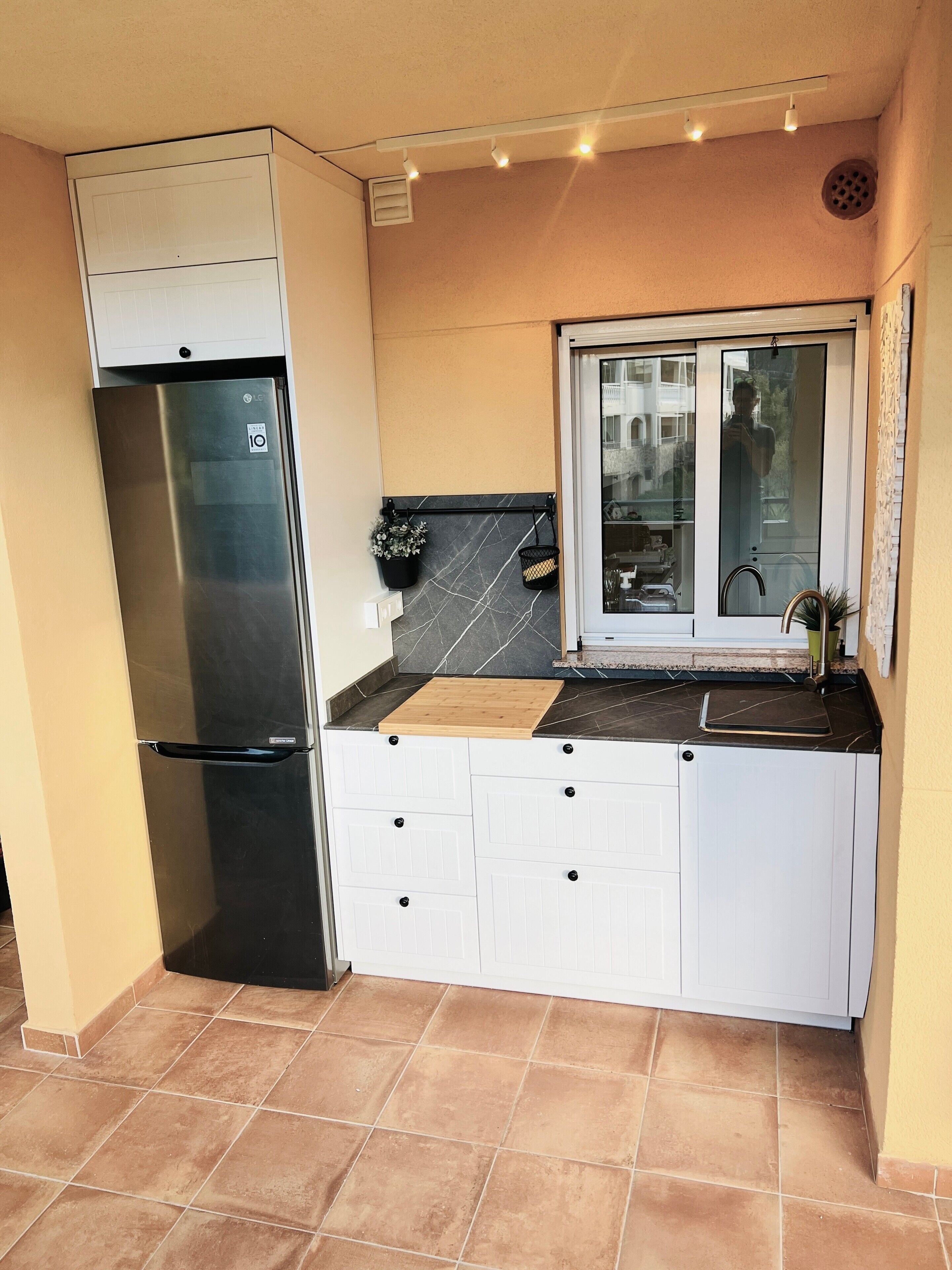 A compact kitchen with a stainless steel refrigerator, white cabinetry, a window, and a tiled floor.