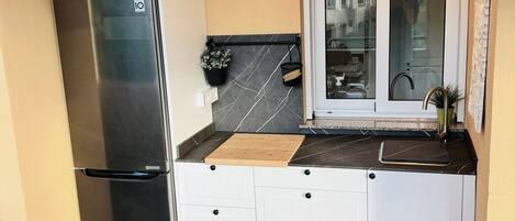 A compact kitchen with a stainless steel refrigerator, white cabinetry, a window, and a tiled floor.