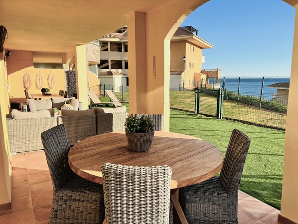 Ein überdachter Außenbereich mit Korbmöbeln, einem runden Holztisch und Blick aufs Meer.