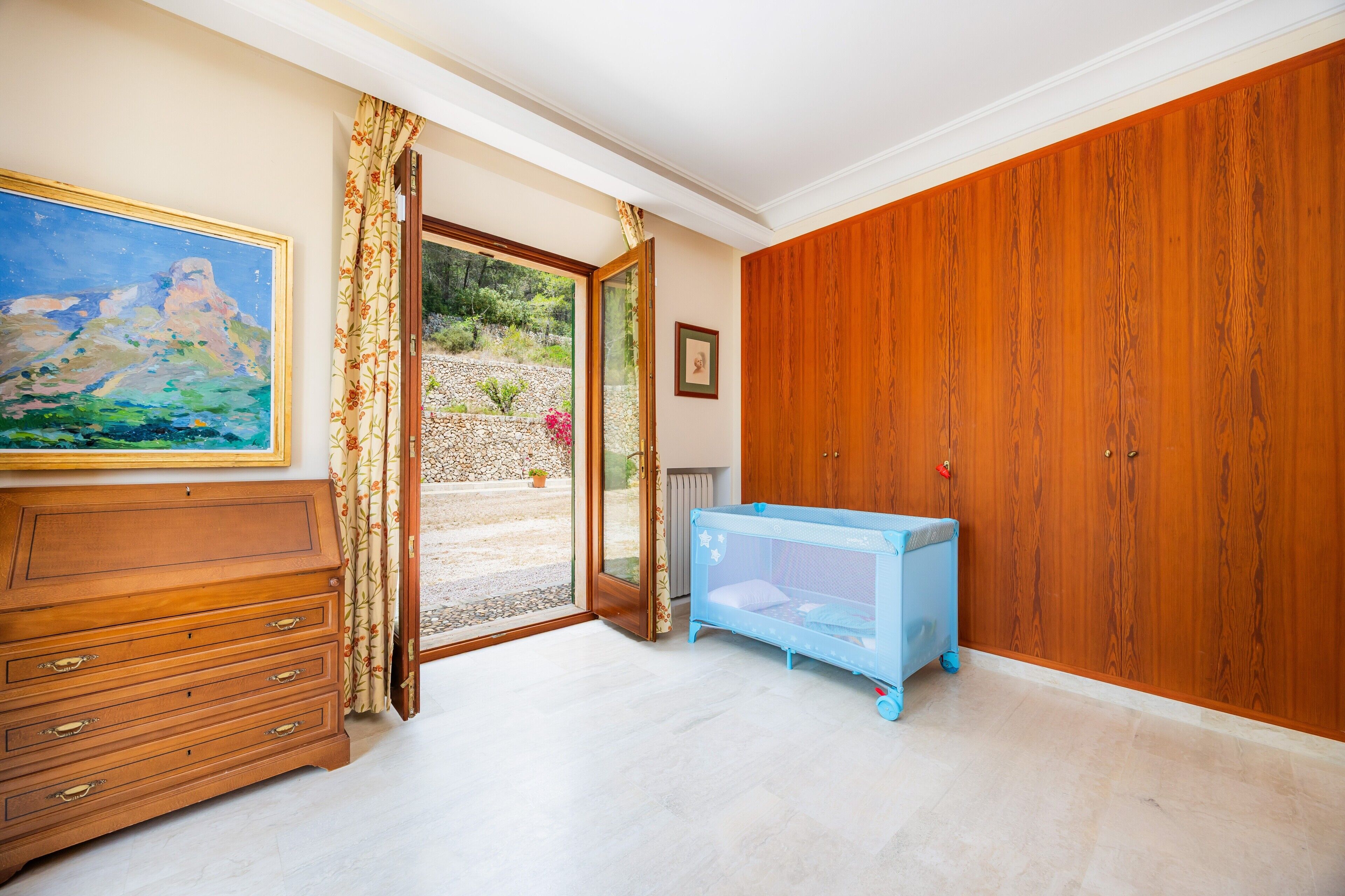 A room with a wooden dresser, a blue crib, a painting, and a view through a glass door.