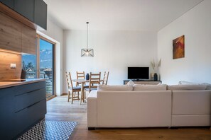 A modern living room with a sofa, TV, dining table, chairs, and a kitchen area with cabinets and a window.