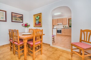 Un coin repas avec des chaises en bois et une table, une cuisine visible à travers une arche, et des œuvres d’art encadrées accrochées aux murs.