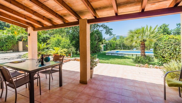 A covered patio with a table and chairs, a potted plant, and a view of a swimming pool and greenery. - Holiday Home 'Sontuiri Den Jaume' with Private Pool, Wi-Fi and Air Conditioning (Pollença)