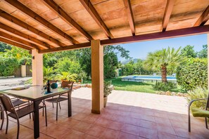 A covered patio with a table and chairs, a potted plant, and a view of a swimming pool and greenery. - Holiday Home 'Sontuiri Den Jaume' with Private Pool, Wi-Fi and Air Conditioning (Pollença)