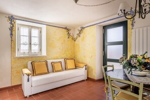 Ein Zimmer mit einer weißen Couch, einem Esstisch mit Stühlen, einem Fenster mit Vorhängen und einer Kristallleuchter.