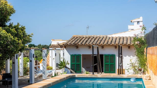A swimming pool with a tiled deck, a white building with green shutters, and a clear blue sky.