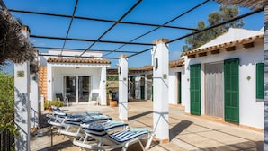 A covered outdoor area with lounge chairs, a patio table, and a building with white walls and green shutters.