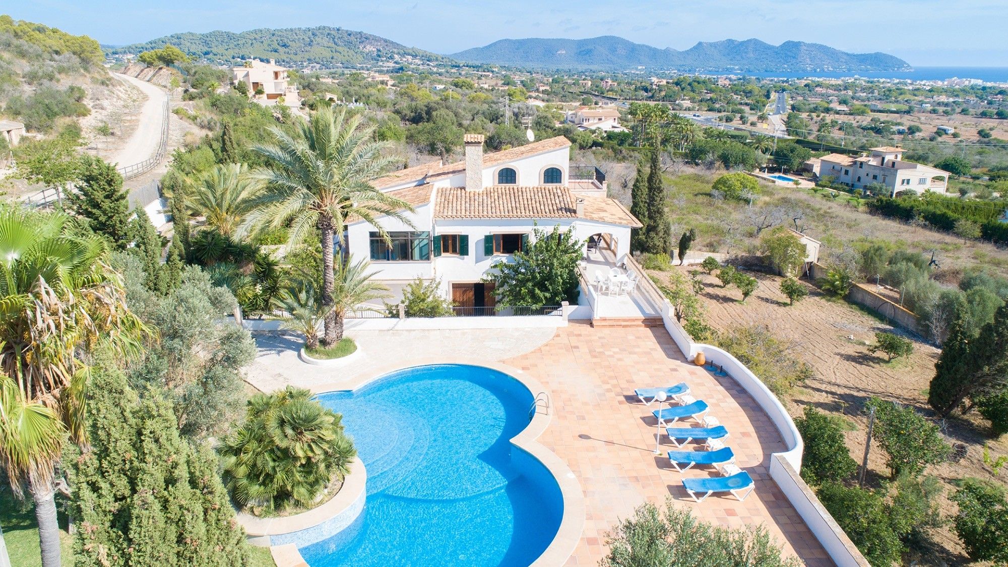 A villa with a swimming pool and sun loungers, surrounded by greenery and a view of the sea.