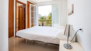 A bedroom with a bed, a bedside lamp, a window with a view of buildings and greenery, and a door. - Minimal Apartment Ref 9000 with Sea View, Balcony, Wi-Fi and Air Conditioning (Can Picafort)