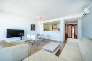 A modern living room with a flat-screen TV, a glass dining table, white sofas, and a kitchen in the background.