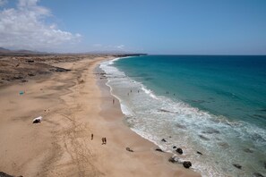 A beach with clear skies, a sandy shore, and a few people walking along the water. - Vacation Home "Casa La Bubilla en Lajares" with Private Garden & Wi-Fi (Lajares)