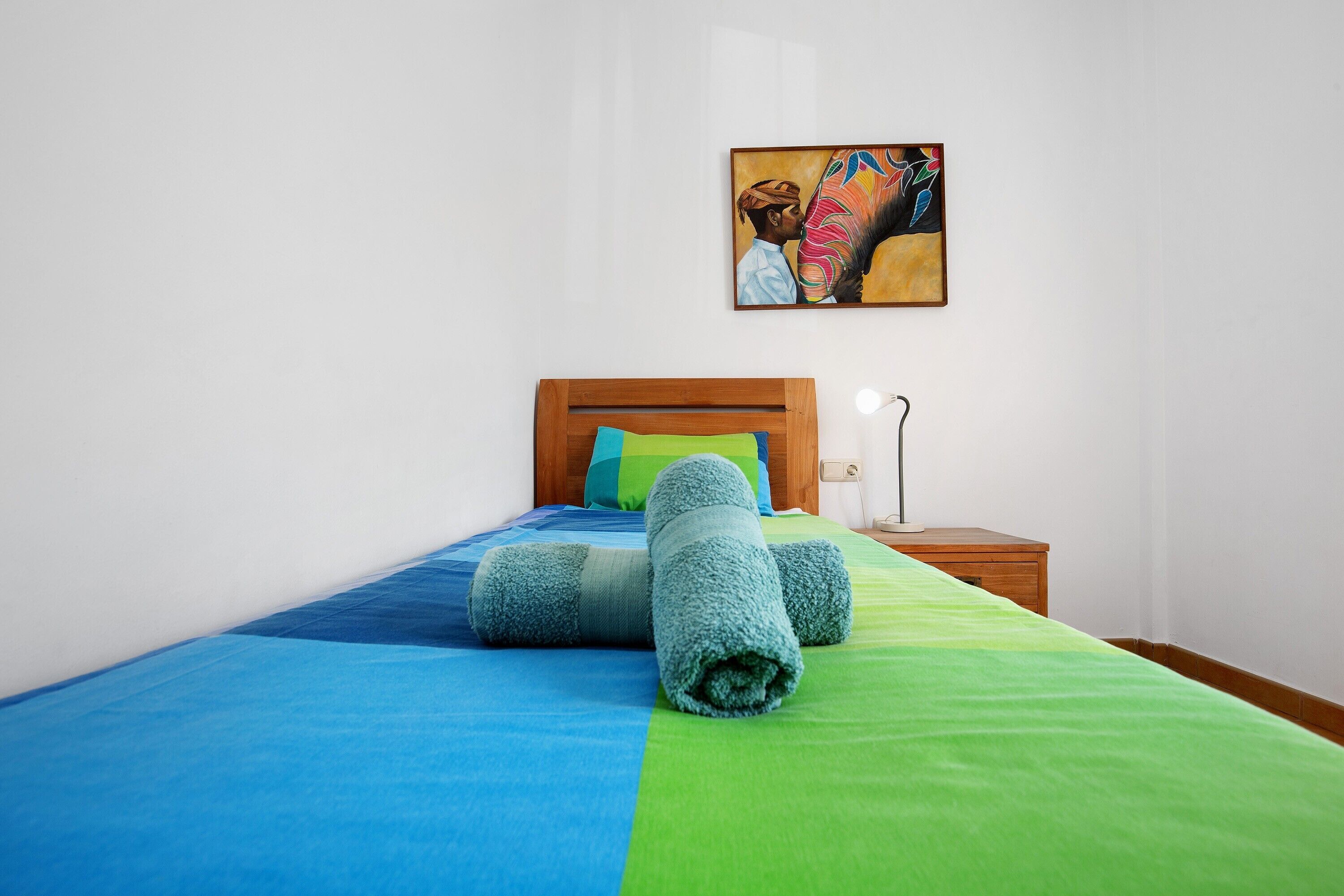 A neatly made bed with blue and green bedding, a wooden headboard, a nightstand with a lamp, and a painting on the wall.