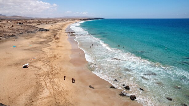 Une plage aux eaux turquoise et limpides, un rivage sablonneux et des montagnes qui se dessinent à l’horizon.
