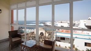 Un balcon avec des fauteuils en osier et une table en verre, offrant une vue sur le paysage urbain de la côte, avec ses bâtiments et la mer.