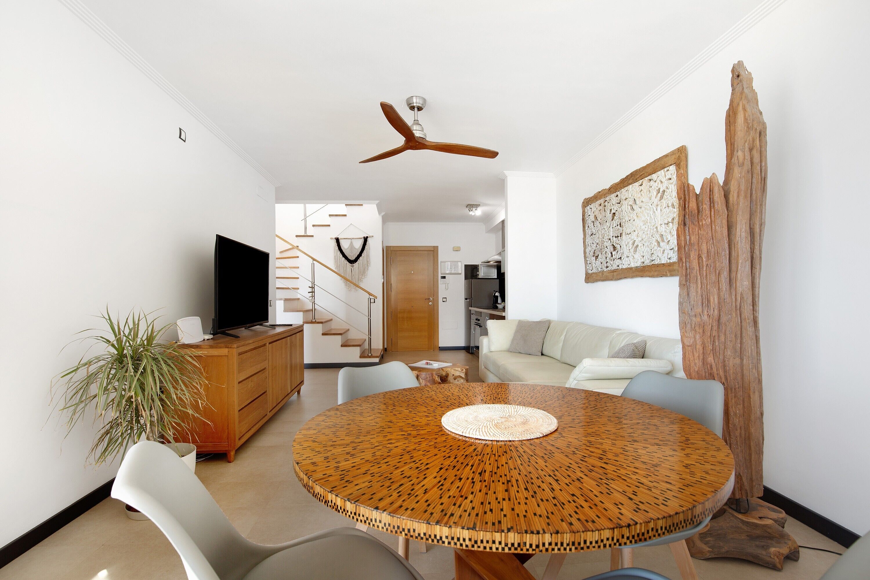 Un salon moderne avec une table ronde en bois, un ventilateur de plafond, un téléviseur, un escalier et un coin repas.