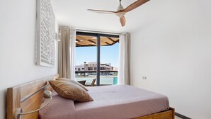 Un dormitorio con cama de madera, ventilador de techo, vista de un edificio a través de la ventana y una silla junto a la ventana.