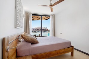 Une chambre avec un lit en bois, un ventilateur de plafond, une vue sur un immeuble par la fenêtre et une chaise près de la fenêtre.