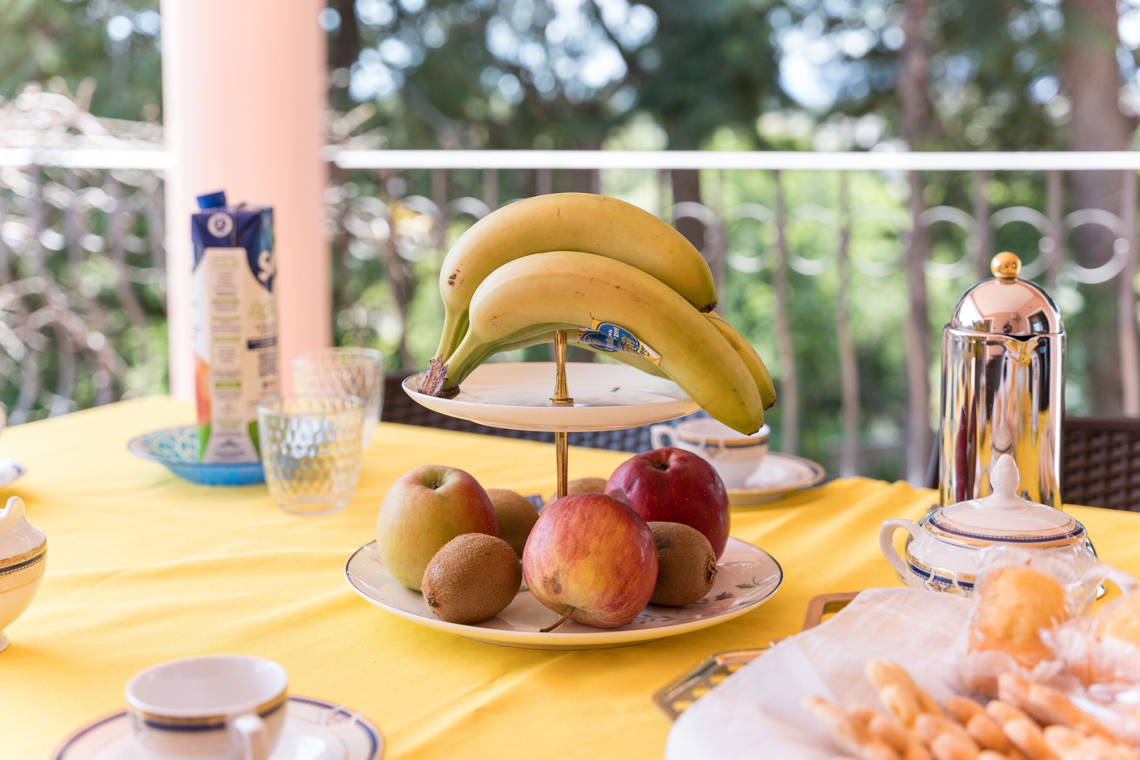 Une table recouverte d’une nappe jaune, une assiette de fruits contenant des bananes, des pommes et des kiwis, une théière et un pichet en verre.
