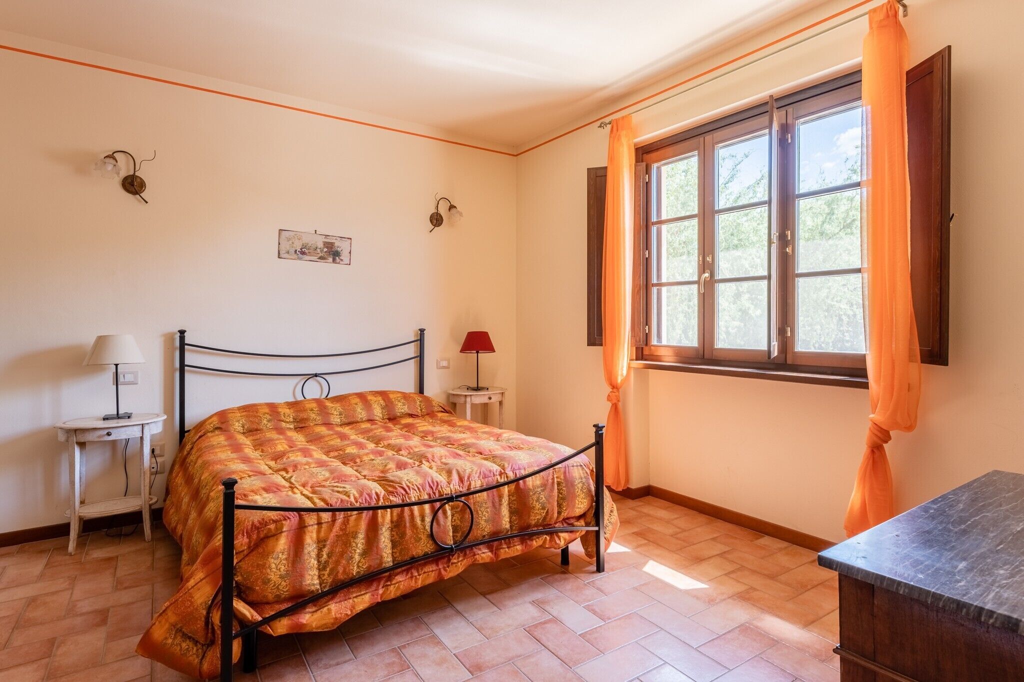 A bedroom with a bed, a nightstand, a lamp, a window with curtains, and a wall-mounted light fixture.