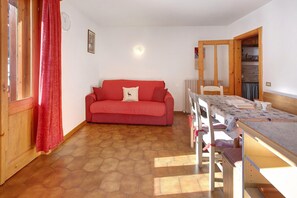 Une pièce avec un canapé rouge, une table à manger en bois recouverte d’une nappe à motifs, un radiateur blanc et une porte en bois.