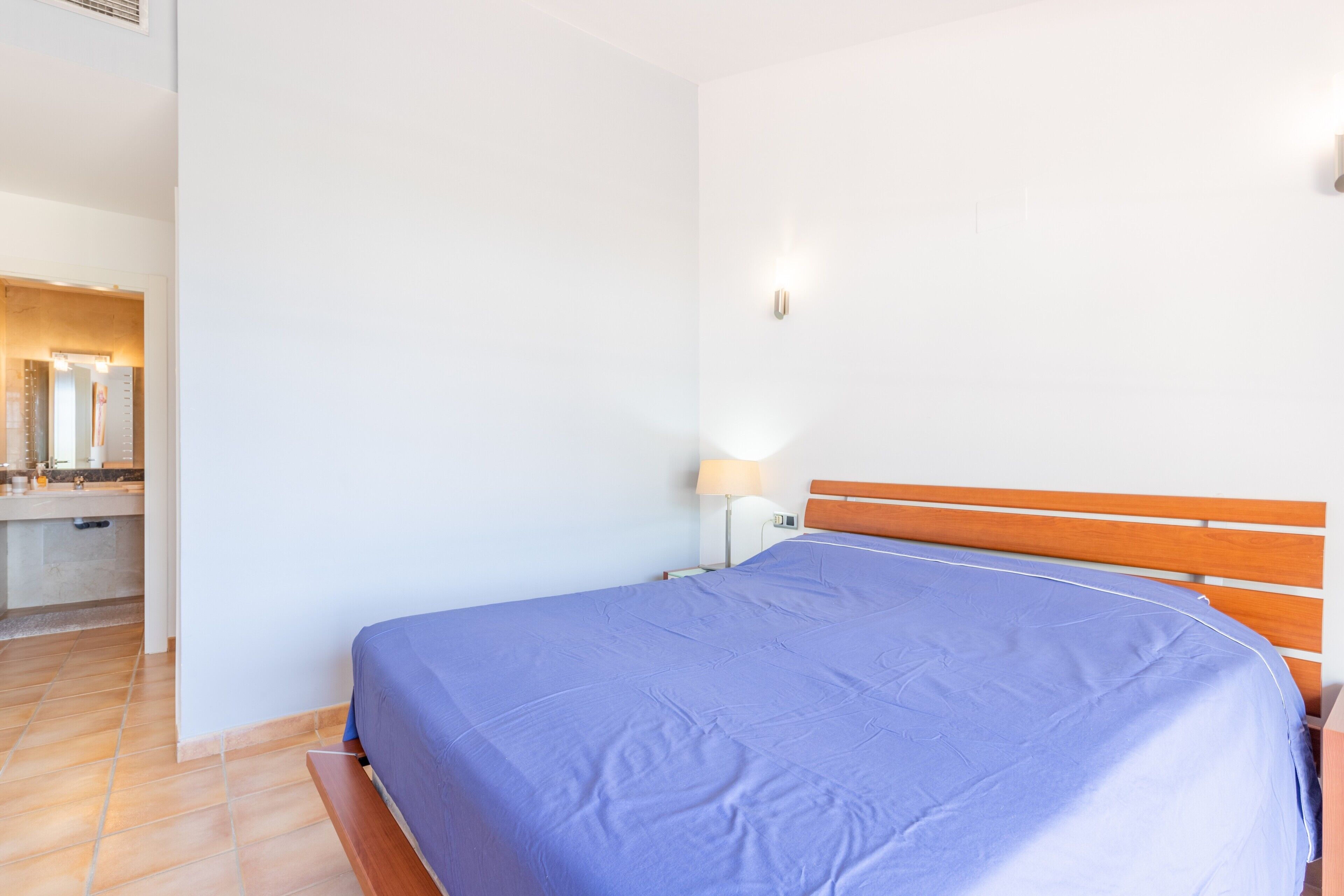 Een slaapkamer met een houten hoofdbord, een bed met een blauwe spreideken, een nachtkastje met een lamp, en een badkamer in de achtergrond.