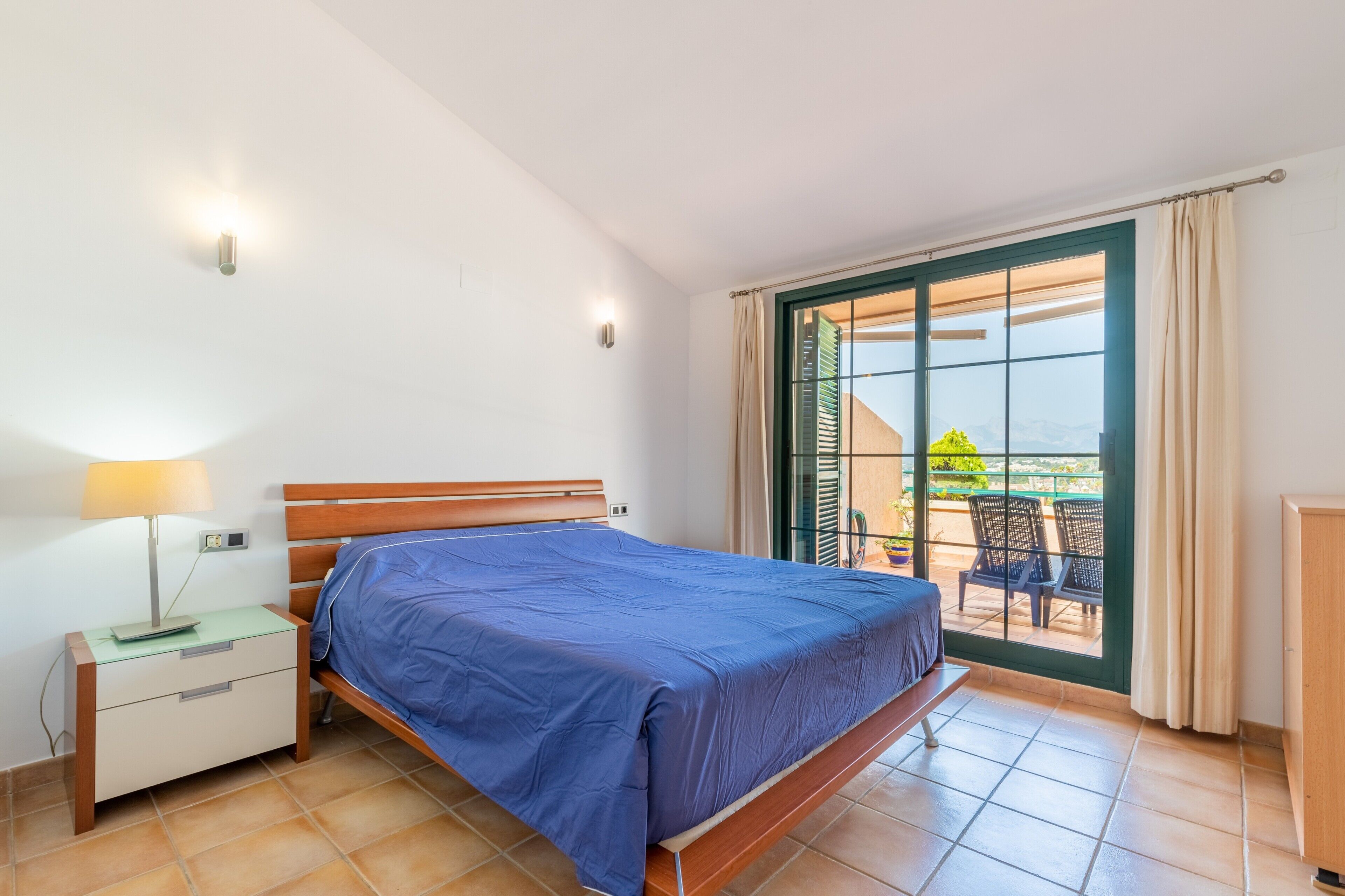 A bedroom with a bed, bedside table, lamp, and a view of an outdoor area with chairs.