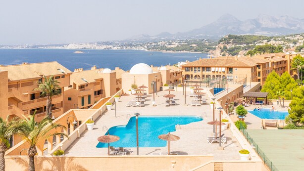 A resort with a pool, sun loungers, umbrellas, and a view of the sea and mountains.
