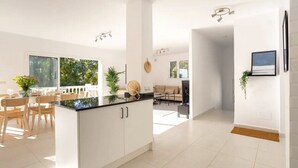 A modern kitchen with white cabinetry, a dining area with wooden chairs, and a view of greenery outside. - Holiday Home 'Modern Villa with Private Pool' with Sea View, Wi-Fi and Air Conditioning (MIJAS)