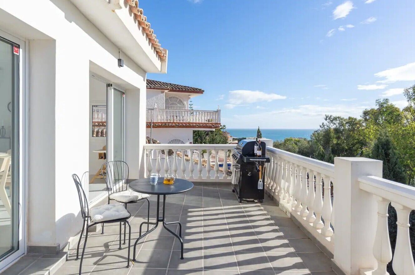A balcony with a table and chairs, a grill, and a view of the sea.