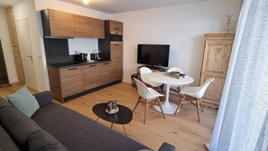 Moderni olohuone, jossa on sohva, ruokapöytä tuoleineen, keittiöalue puukuvioisilla kaapeilla ja televisio.