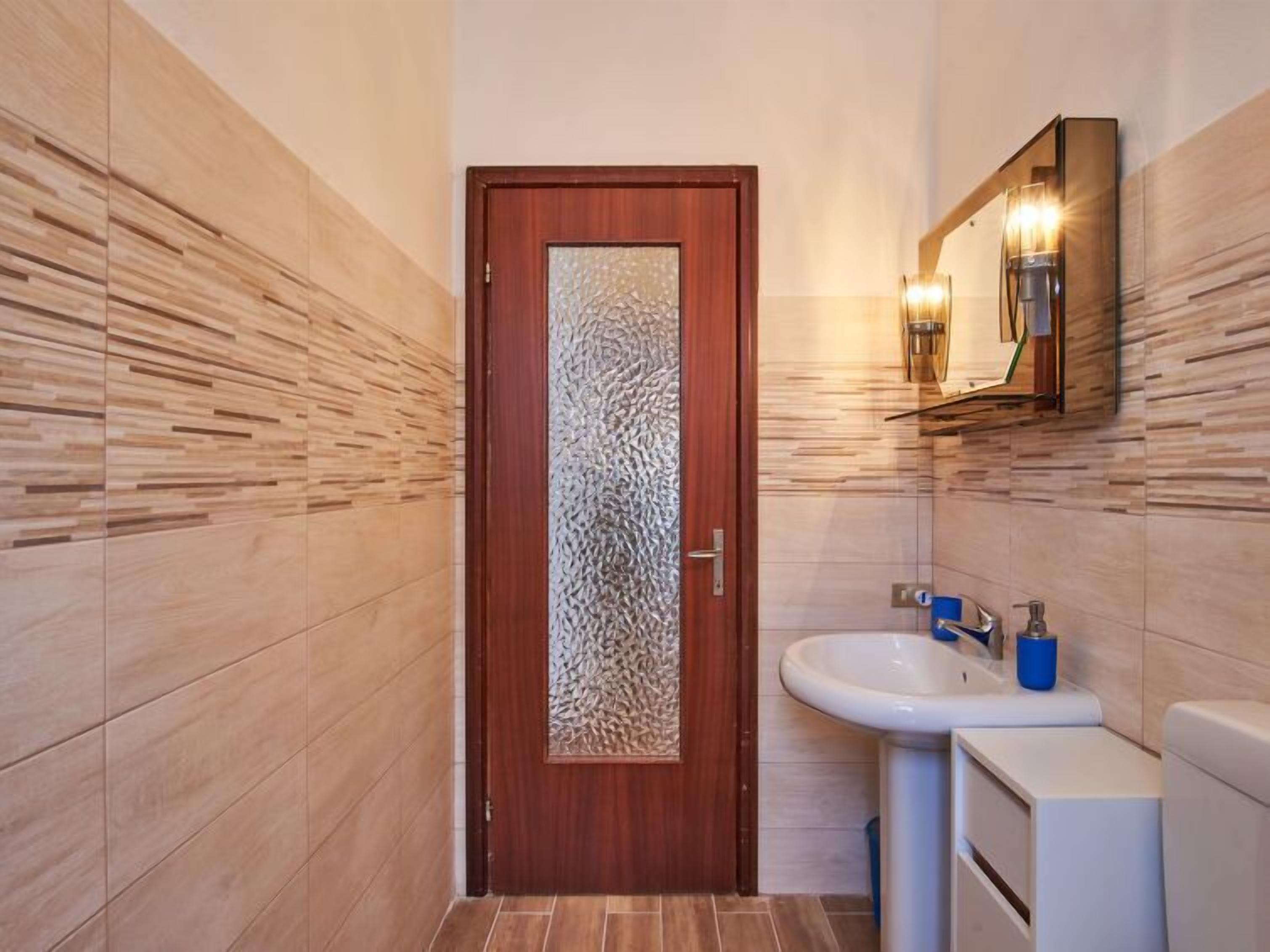 A bathroom with a wooden door, a sink with a mirror, and a wall-mounted light fixture.