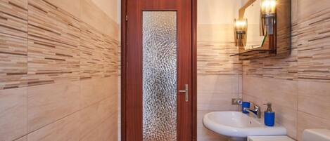 A bathroom with a wooden door, a sink with a mirror, and a wall-mounted light fixture.