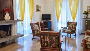 A living room with a fireplace, two wooden chairs with floral upholstery, a television, and yellow curtains.