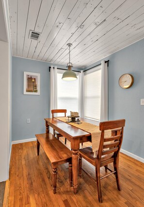 A dining area with a wooden table and benches, a hanging light fixture, a framed picture on the wall, and a window with curtains.