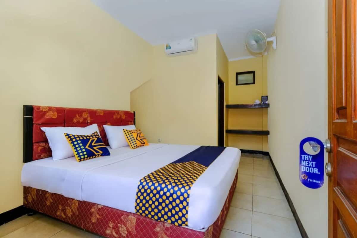 Une chambre d’hôtel avec un lit, une tête de lit rouge, une couvre-lit blanche et bleue, et une pancarte sur la porte.