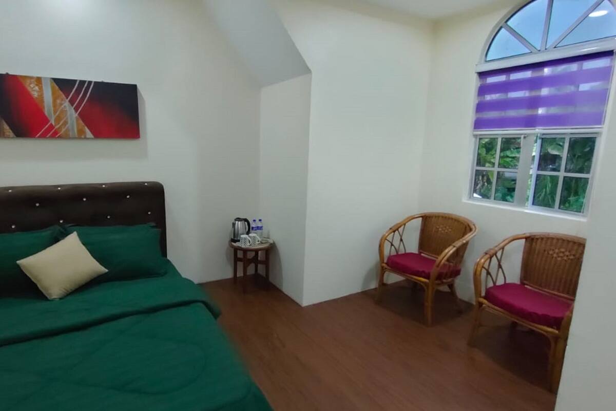 A bedroom with a bed, two wicker chairs, a small table with a kettle, and a window with a view of greenery.