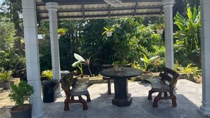 Een overdekt terras met een tafel en twee stoelen, omgeven door potplanten en een pergola.